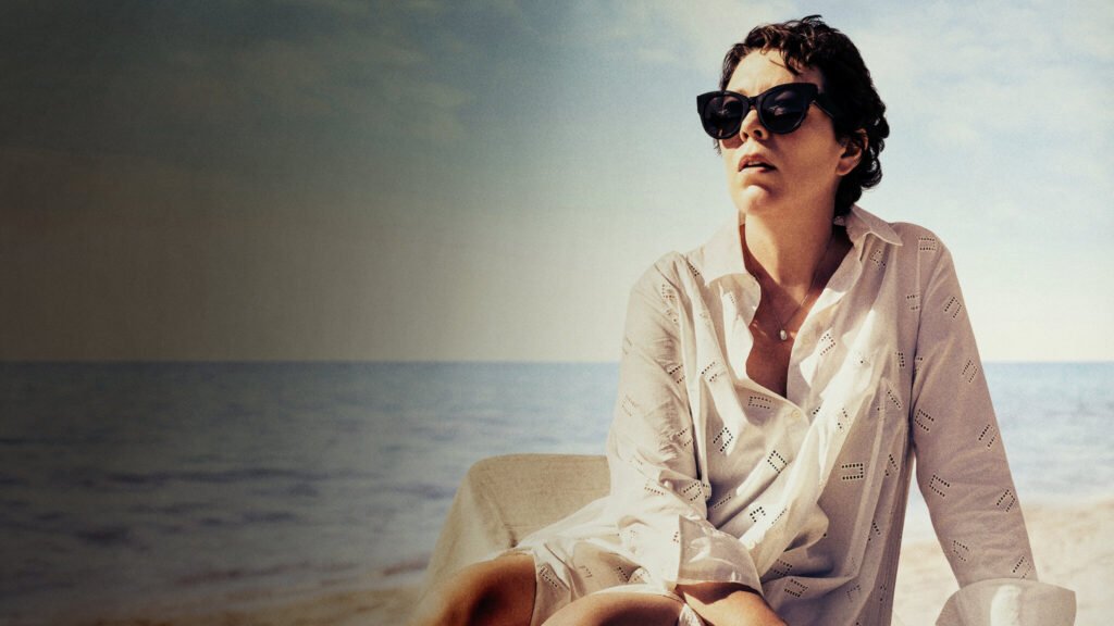 Mulher sentada à beira-mar, usando óculos escuros e camisa clara, em um clima introspectivo que representa o tom dramático do filme A Filha Perdida, um dos melhores filmes originais Netflix com foco psicológico.