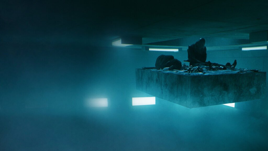 Plataforma de concreto descendo em um ambiente minimalista e sombrio, com dois prisioneiros recolhendo restos de comida — cena marcante do filme O Poço, um dos melhores filmes originais Netflix e um dos mais comentados do catálogo.