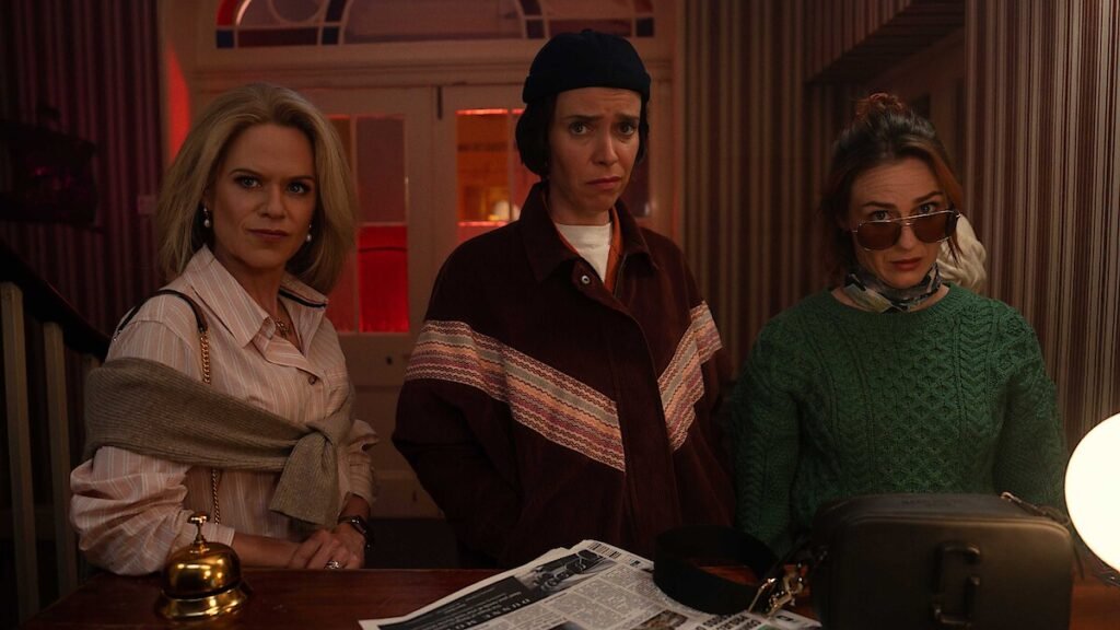 Imagem da série How To Get To Heaven From Belfast (2026), mostrando três mulheres no balcão de um hotel — uma elegante e confiante, outra com expressão de preocupação e a terceira usando óculos escuros e visual excêntrico. A cena tem iluminação quente e clima de comédia misteriosa, representando um dos destaques entre os lançamentos Netflix 2026.