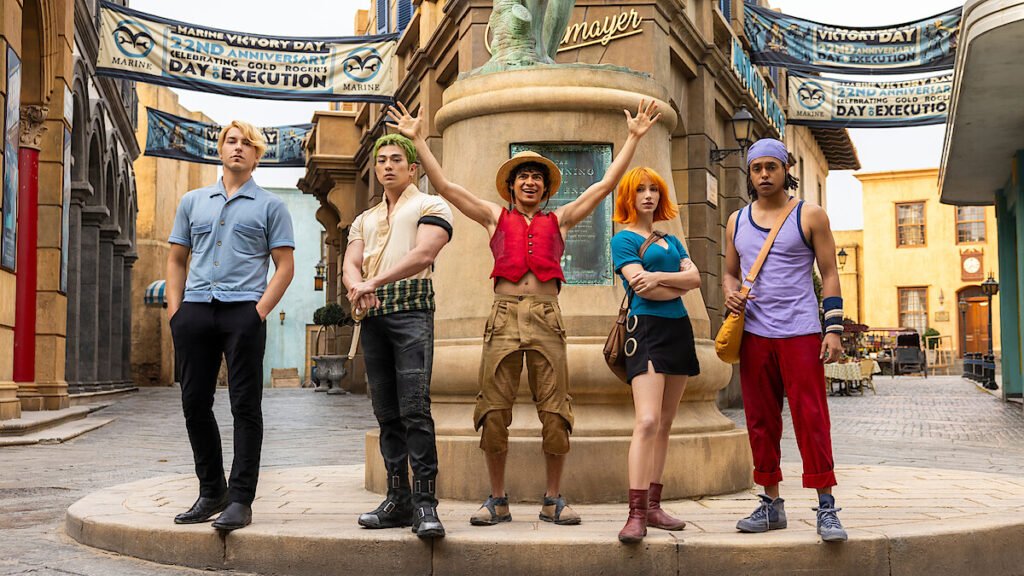 Imagem promocional de One Piece – Temporada 2 (Live Action) mostrando os cinco protagonistas — Sanji, Zoro, Luffy, Nami e Usopp — posando em uma rua temática, com bandeiras da Marinha ao fundo. A equipe aparece com figurinos fiéis ao mangá e vibe de aventura épica, destacando a série como um dos principais lançamentos Netflix 2026.