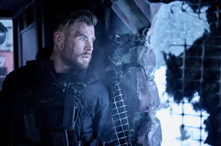 Chris Hemsworth como Tyler Rake em cena de ação do filme Resgate 3 da Netflix, segurando uma arma e com expressão de cansaço em meio ao combate.
