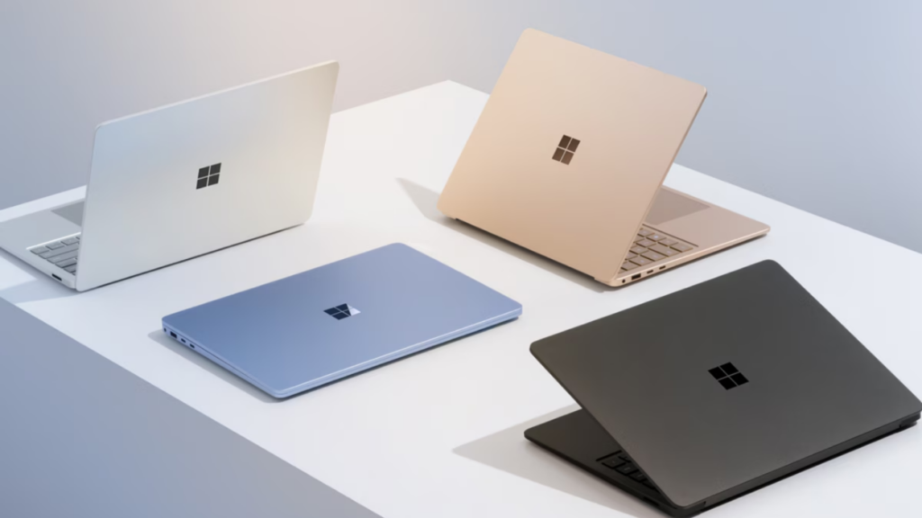 Linha de notebooks Microsoft Surface em diferentes cores — branco, azul, dourado e preto — exibidos sobre uma mesa minimalista, destacando o design premium da marca.